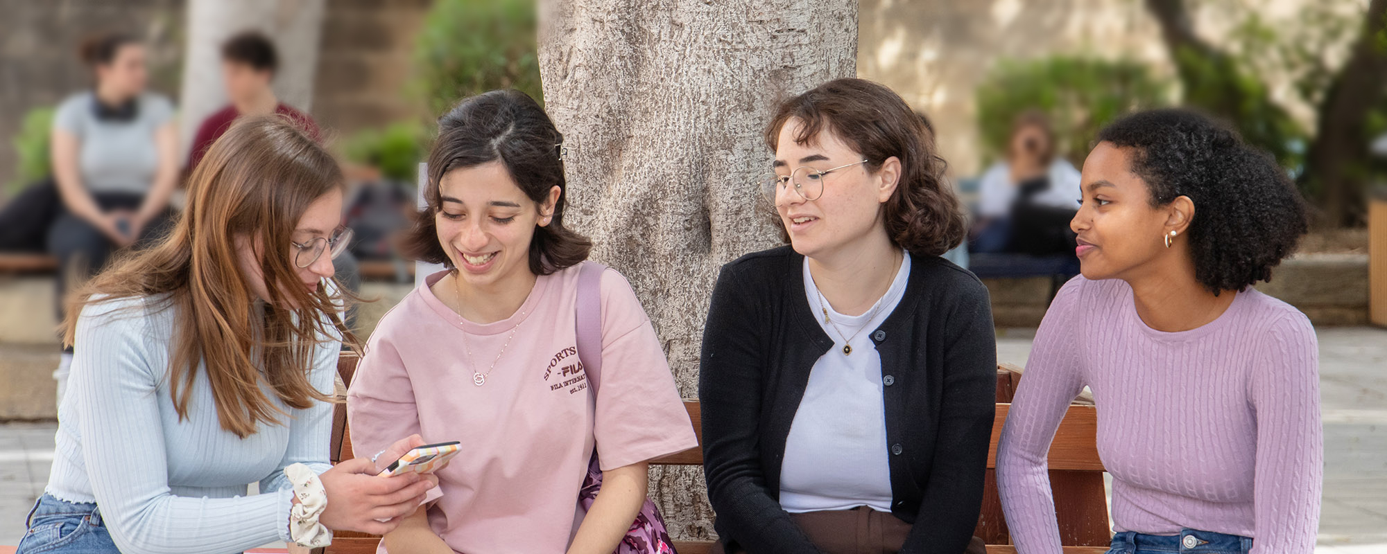 Four girls sitting in an outdoor space with a tree behind them; one of the girls has a smartphone in her hand