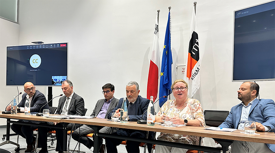 Speakers at a conference sitting at a table with flags behind them